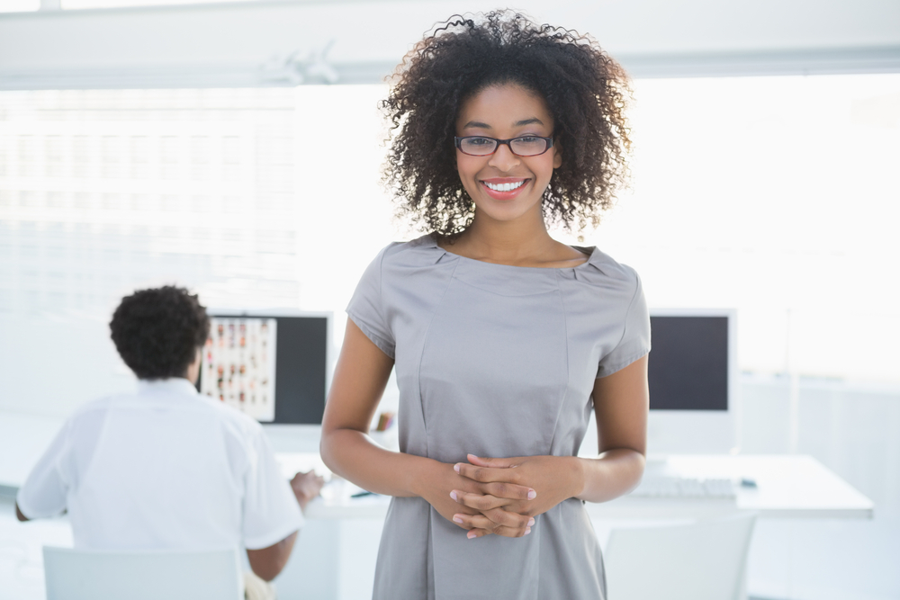 Young pretty editor smiling at camera with colleague working behind her in creative office Young pretty editor smiling at camera with colleague working behind her in creative office